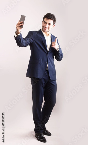 Obraz Handsome bearded young business man taking selfie smiling. Full body length portrait isolated over grey studio background.