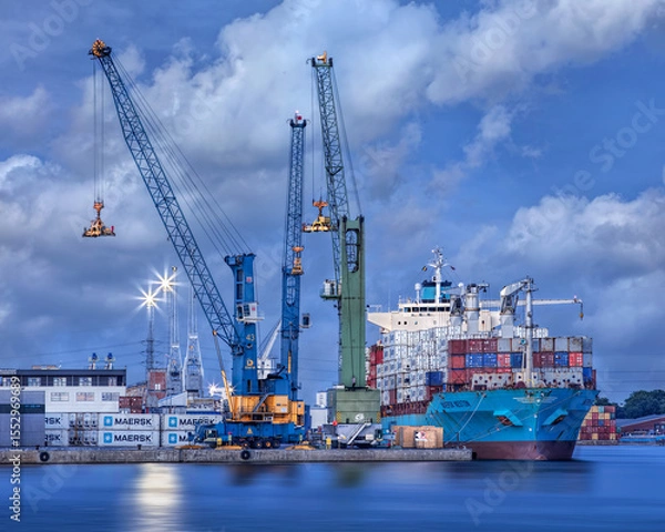 Obraz Container ship moored at Port of Antwerp during twilight with large cranes and dramatic clouds.
