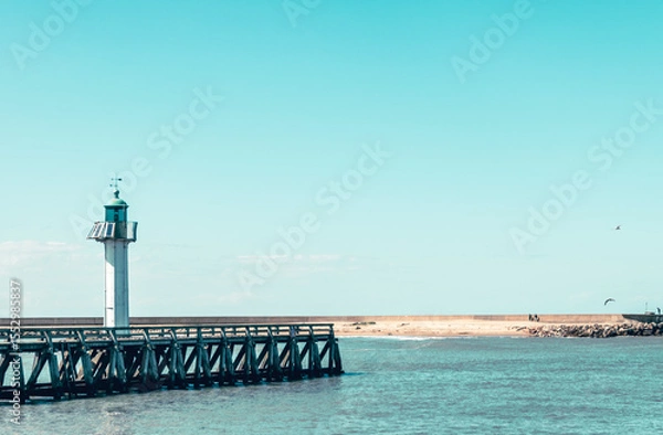 Fototapeta Scenic view of a lighthouse near a wooden pier under clear blue skies in a coastal location