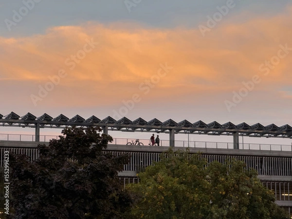 Fototapeta Silhouetted pedestrians with bicycle traverse an elevated modern walkway beneath a dramatic orange sunset sky framed by angular metal canopy structures above leafy trees