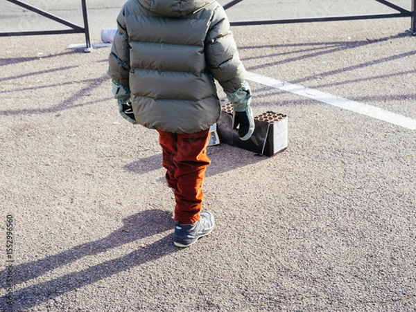 Fototapeta Child in winter clothing stands in a parking lot observing a used fireworks box in daylight, possibly after a New Year celebration event.