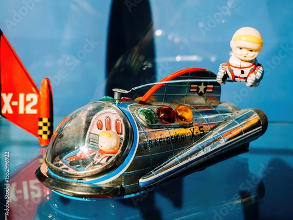 Fototapeta Colorful vintage tin space toy rocket labeled X-12 with astronauts in suits displayed in a museum setting against a vibrant blue background
