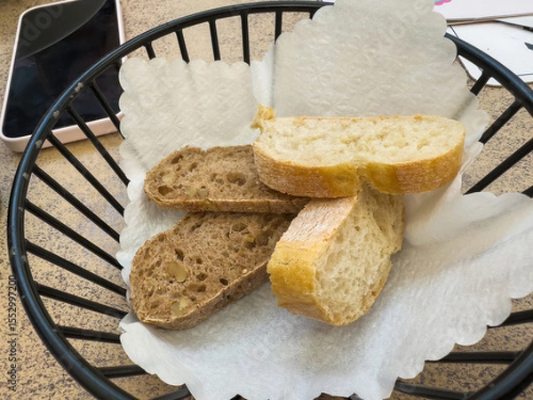 Fototapeta A black wire basket lined with white paper holds slices of brown walnut bread and white crusty bread on a restaurant table next to a smartphone and paper coasters