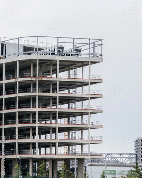 Fototapeta Exposed concrete skeleton of a multi story building under construction featuring scaffolded levels safety railings and heavy machinery set against Strasbourg skyline
