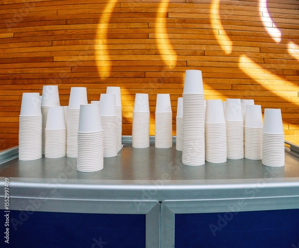 Fototapeta Neatly stacked white paper cups arranged on a metal counter in a modern conference room with warm wooden wall panels and artistic lighting.