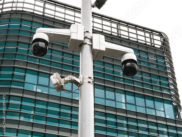 Fototapeta Paris, France - May 4, 2025: Modern surveillance cameras mounted on a pole in front of a curved glass office building, symbolizing urban security and monitoring technology