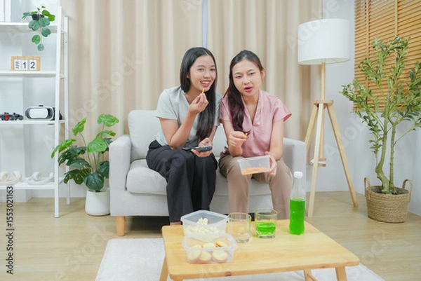 Fototapeta Happy friends asian woman enjoying watching television with popcorn using remote control choosing TV show movie. Enjoying television relaxing at home together.