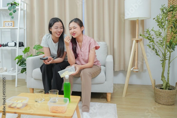 Fototapeta Happy friends asian woman enjoying watching television with popcorn using remote control choosing TV show movie. Enjoying television relaxing at home together.