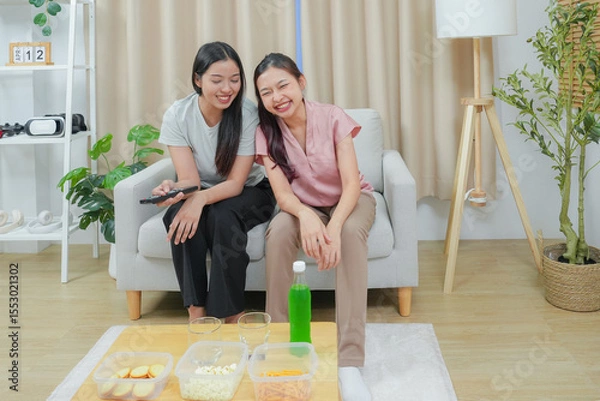Fototapeta Happy friends asian woman enjoying watching television with popcorn using remote control choosing TV show movie. Enjoying television relaxing at home together.