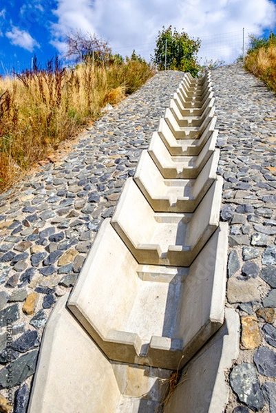 Fototapeta Concrete drainage channel on a hillside lined with stones surrounded by dry grass and shrubs under a blue sky