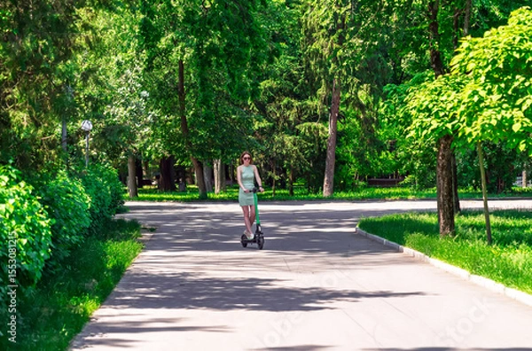 Obraz Modern urban transport: A stylish girl in a dress enjoying a ride on an e-scooter in nature.Summer leisure and active lifestyle. A woman on a kick scooter in a public city park.Eco-friendly commute