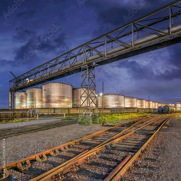 Obraz Industrial scene whit rails on the foreground, shiny silos on the background and al large pipeline bridge, Port of Antwerp, Belgium