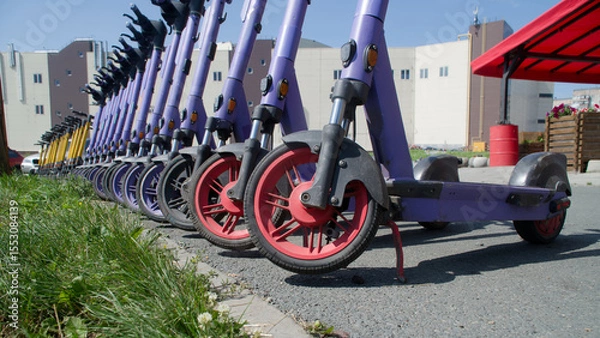 Obraz A line of purple and yellow electric scooters parked on a city sidewalk, highlighting modern micro-mobility services in an urban environment. Photo. Selective focusing