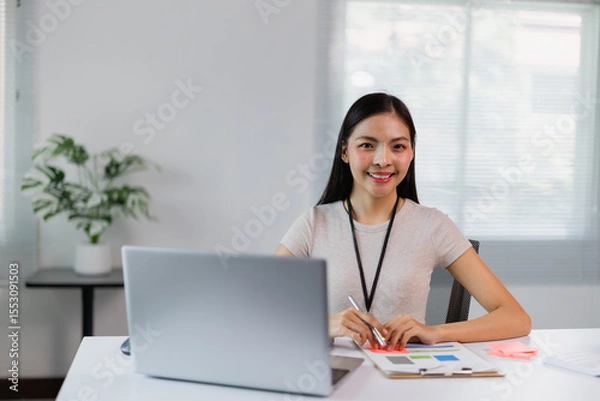 Fototapeta Young Asian businesswoman analyzing financial data on a laptop while taking notes on a clipboard, working diligently at her modern office desk with natural light streaming in
