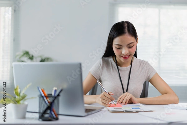 Fototapeta Young Asian businesswoman smiling and working with financial documents using laptop and taking notes on a sticky note while sitting at her desk in a modern office