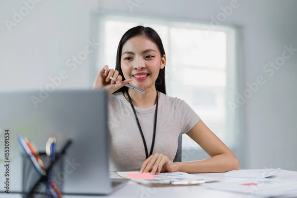 Fototapeta Young asian businesswoman working with laptop and holding pen while sitting at her desk in modern office, smiling and looking towards the laptop