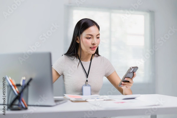 Fototapeta Young office employee wearing an ID card, using a smartphone while working on a laptop and managing documents on a desk in a modern, well-lit office environment