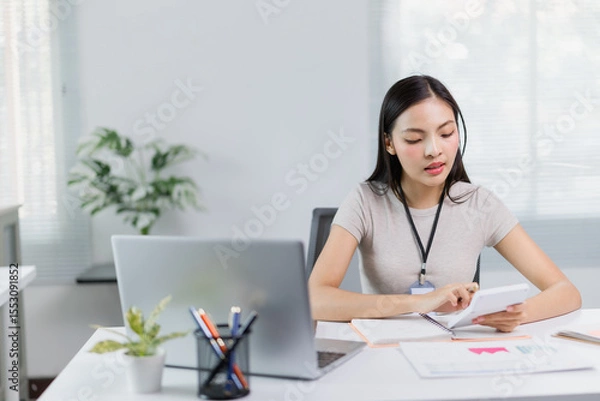 Fototapeta Young focused asian businesswoman wearing headset using calculator and working with laptop computer at modern office, business and finance concept