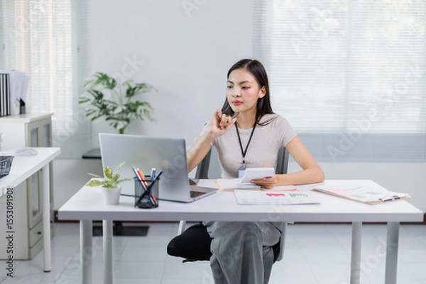 Fototapeta Young office worker sitting at desk and using laptop while holding calculator and reviewing documents, performing calculations and analyzing financial data for business strategy