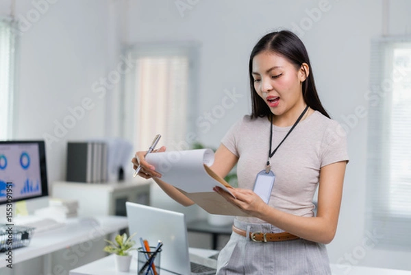 Fototapeta Young Asian businesswoman wearing an ID card is standing and reviewing documents while preparing notes in a modern office with computers and charts on the screen
