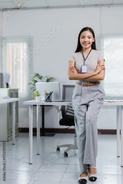 Fototapeta Young Asian businesswoman wearing identification card is standing with arms crossed and smiling in modern office, showing confidence and professionalism
