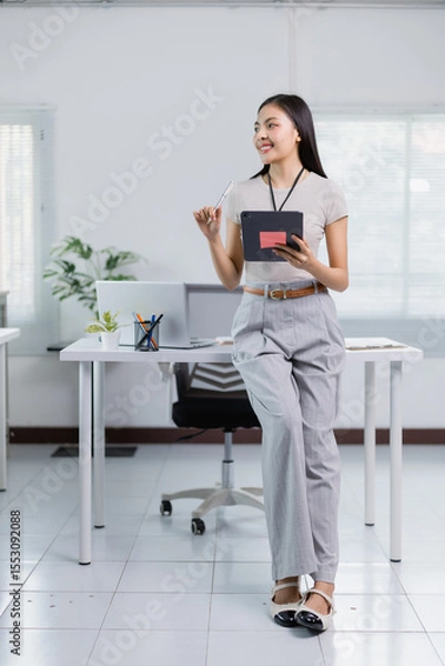 Fototapeta Young businesswoman is holding a tablet and a pen, looking away and thinking about new ideas, leaning against a desk in a modern office, smiling and confident in her work