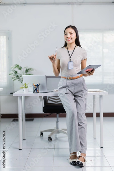Fototapeta Young businesswoman is holding a tablet and a pen while having a new idea, leaning on her desk in a modern office, smiling and looking away