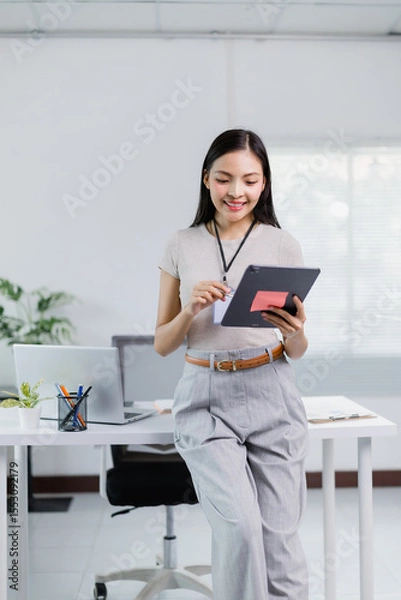 Fototapeta Young Asian businesswoman wearing lanyard with id card and using tablet, leaning on desk in modern office with laptop, smiling and looking at screen
