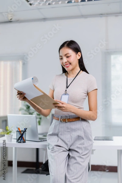 Fototapeta Smiling businesswoman wearing an ID card stands in her office, reviewing documents while a laptop and a neatly organized desk provide a modern backdrop to her professional environment