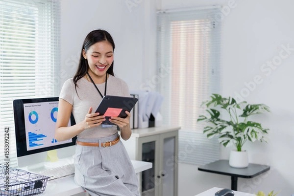 Fototapeta Young office worker holding digital tablet and checking project statistics on computer, enjoying work in modern company office with big windows and plants