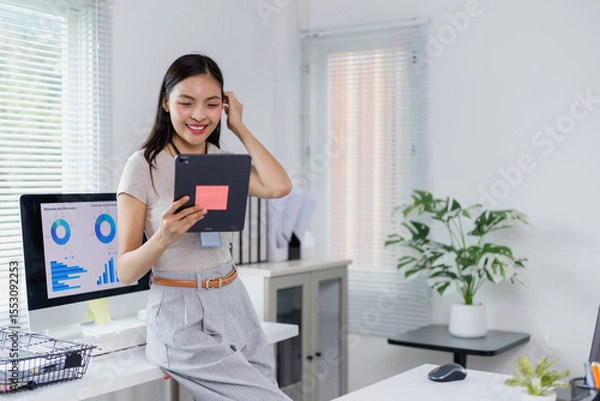 Fototapeta Businesswoman is using a tablet and analyzing financial data on a computer in the office, touching her hair with a thoughtful expression as she reviews the information on the screen