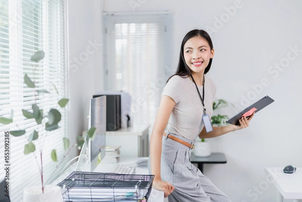 Fototapeta Young Asian businesswoman wearing gray trousers and beige t-shirt, holding tablet and smiling while leaning on desk in modern office with computer and blinds on window