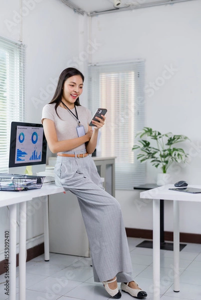 Fototapeta Young businesswoman wearing an id card and leaning on her desk while using her smartphone in a modern office, with a computer showing charts