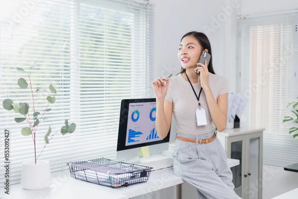 Fototapeta Young businesswoman wearing an id card is sitting on her desk and talking on the phone while holding a pen and looking away, with computer showing graphs in front of her in a modern office