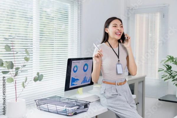 Fototapeta Young office worker happily talking on the phone and holding a pen while sitting on her desk, with a desktop computer displaying charts and graphs in the background, suggesting a productive workday