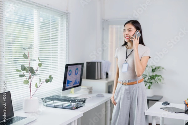 Fototapeta Young Asian businesswoman wearing an ID card, talking on phone standing in modern office with computer and laptop on desk, looking away and smiling