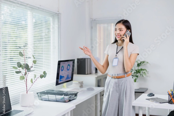 Fototapeta Businesswoman wearing an ID card is standing in a bright office, talking on her smartphone and pointing at a computer screen displaying charts, suggesting a discussion about business performance