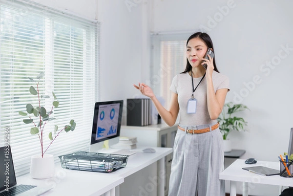 Fototapeta Businesswoman is standing in a bright, modern office, talking on her phone and gesturing with her hand, suggesting she's engaged in a discussion or giving instructions