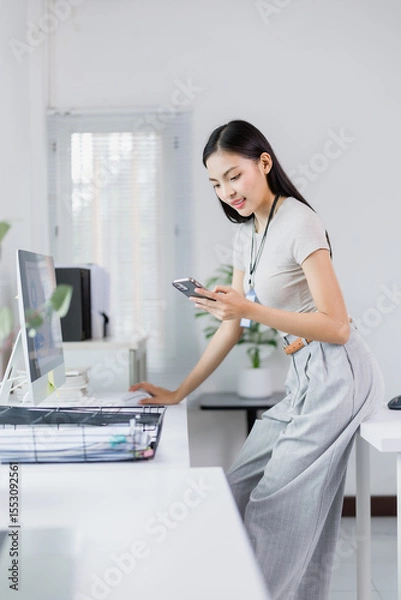 Fototapeta Young Asian businesswoman working on a computer while using a smartphone in a modern office, leaning on her desk and checking messages in a bright, clean environment