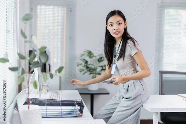 Fototapeta Young Asian businesswoman wearing lanyard with id card is using smartphone and working with desktop computer in modern office, business and technology concept