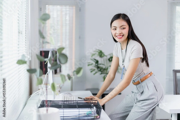 Fototapeta Young Asian businesswoman smiling while leaning on her desk in a modern office workspace with plants and natural light, showcasing a positive and productive work environment