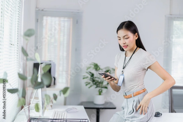 Fototapeta Young Asian businesswoman using mobile phone in modern office with computer and plants, she is wearing casual clothes and standing near her desk