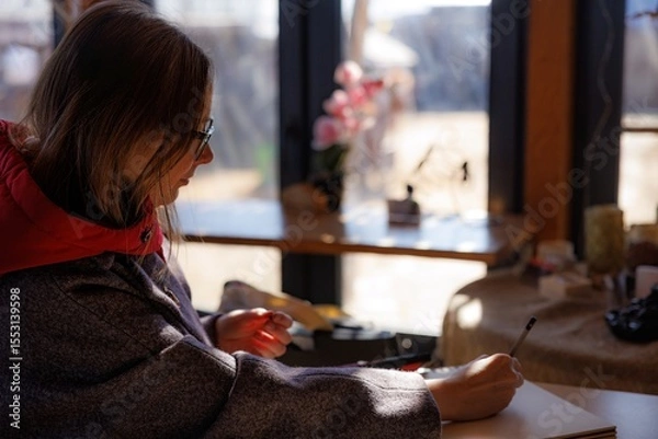 Fototapeta A woman with glasses is deeply focused on sketching at a sunny table in a warm, inviting studio atmosphere.