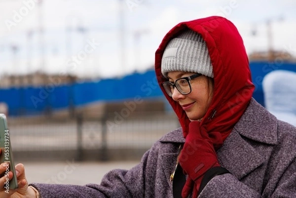 Fototapeta Dressed warmly, a woman enjoys taking a moment to connect with her smartphone while standing outside in the city.