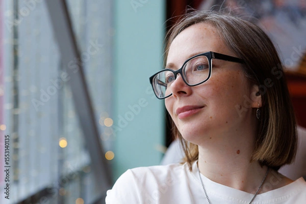 Fototapeta A thoughtful woman gazes outside through the large window of a bright cafe, enjoying a peaceful moment.