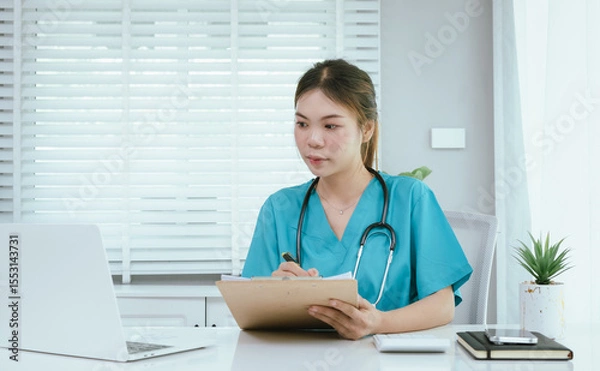 Fototapeta A female medical professional, who appears to be an East Asian female, in turquoise scrubs and a stethoscope, is actively working on a laptop while taking notes.
