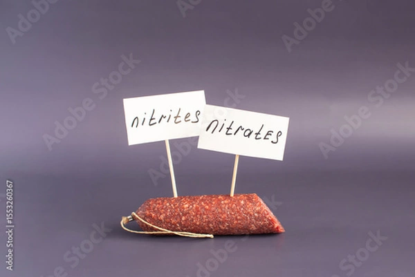 Fototapeta A fragment of smoked sausage and the warning labels "Nitrates and nitrites" in close-up on a dark background. The danger of harmful substances in sausage products