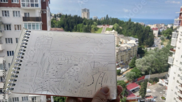 Fototapeta Person is holding a sketchbook with a drawing of a city in the background. The drawing is of a city with a sea on horizon in Sochi