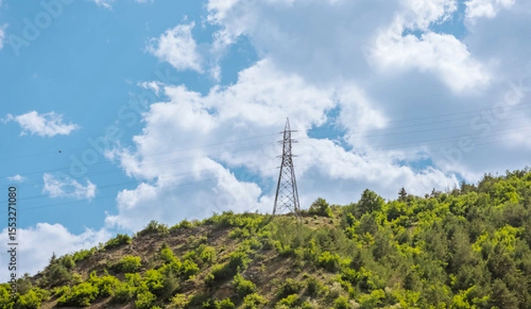 Fototapeta Power line tower on green hill under cloudy sky. This scene captures the intersection of nature and infrastructure. Use for themes related to energy and landscape.