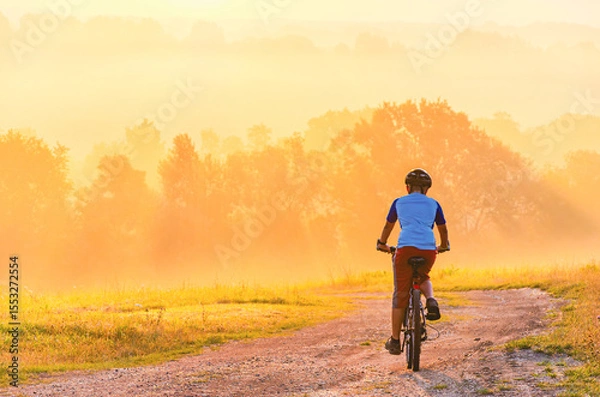 Fototapeta A man is riding a bike on a dirt road. The road is surrounded by trees. The sky is orange and the sun is setting